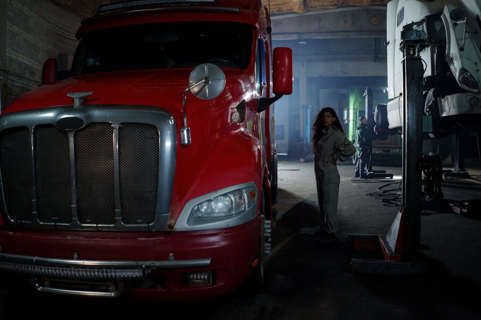 Female mechanic with curly hair stands confidently next to a red truck in a garage.